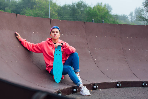 Stylish young woman with short colored hair sitting with her blue plastic skateboard in skatepark. Youth concept. - Foto, imagen