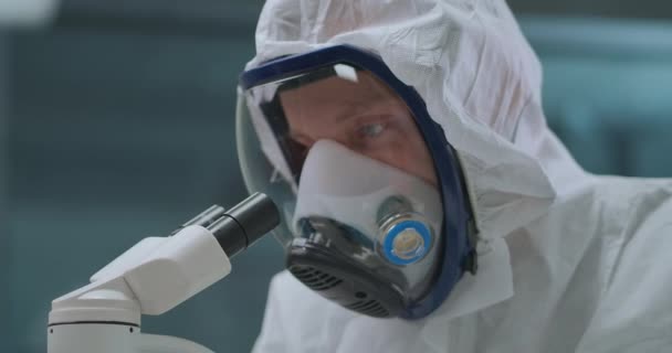 el hombre está utilizando el microscopio electrónico moderno para explorar bacterias y virus en el laboratorio, protegiéndose por máscara con respirador - Metraje, vídeo