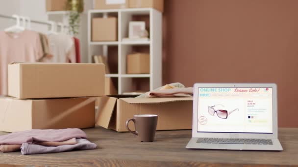 PAN shot of interior of office of small retail company. Cardboard boxes, folded clothes and laptop with online marketplace web page open on it are on table - Footage, Video
