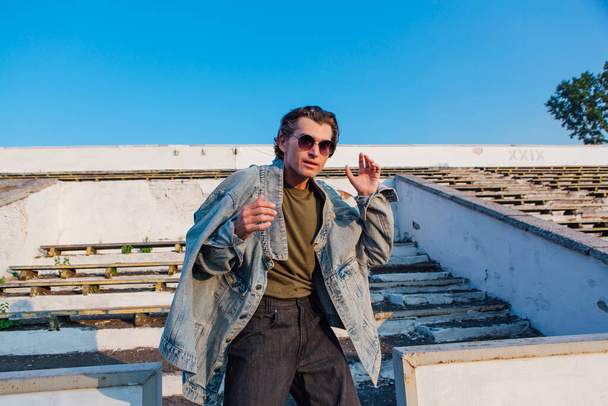 Fashion portrait of a tall handsome man dressed in jeans jacket ans sunglasses on the crushed tribune of the old stadium during sunset - Φωτογραφία, εικόνα