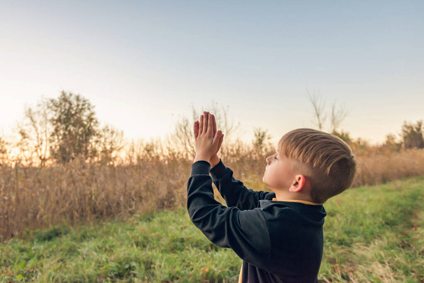 Egy kisgyerek felemeli a kezét az égre, imádkozva az esti parkban. - Fotó, kép