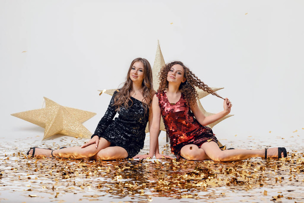 Akşam giyimli iki güzel kadın altın konfetili açık bir arka planda bir partide giyiniyor. Kutlama konsepti, stilin güzelliği. - Fotoğraf, Görsel