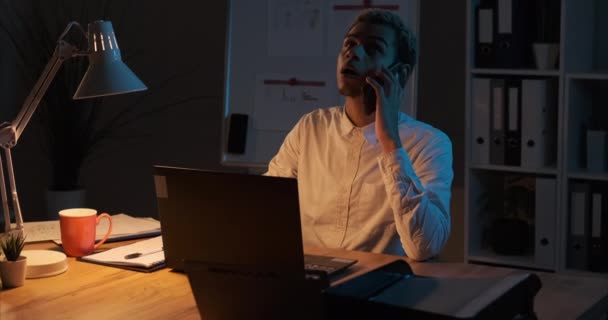 Podnikatel analyzuje dokumenty při používání notebooku a mluví na mobilním telefonu v noční kanceláři - Záběry, video