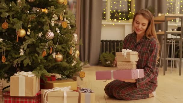美しい装飾されたクリスマスツリーの下でラップに座っているパジャマで笑顔の若い女性の完全なショット真夜中にクリスマスの前夜にプレゼントボックスを置きます - 映像、動画
