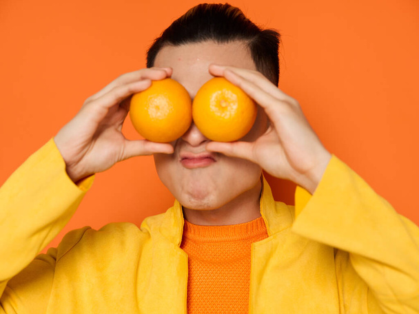 Cheerful man with orange fruit in front of his eyes and in a yellow jacket cropped view - Fotografie, Obrázek