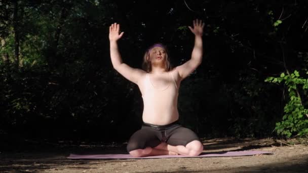 Hravý tlustý muž s dlouhými vlasy v těsné tričko legrační jóga na Fitness Mat. Šílenec zapojený do outdoorového výcviku v lese - Záběry, video