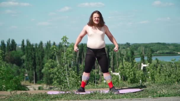 ピンクのTシャツとタイトレギンスで長い髪を持つ遊び心のあるファットマンは、屋外で減量のための体操を行います。公園でダンベルとフィットネスに従事女の子を描いた面白い太りすぎの男.スローモーション - 映像、動画
