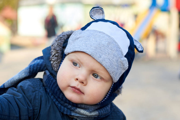 Портрет милого маленького голубоглазого мальчика с интересным взглядом в шляпе, шарфе и куртке на улице ранней весной - Фото, изображение