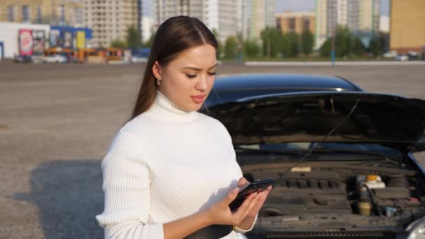 Jonge vrouw aan de telefoon praten tegen de achtergrond van de open motorkap van de auto - Video