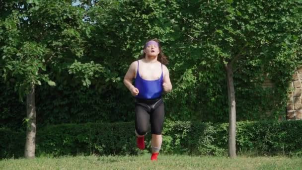 Playful Fat Man with Long Hair in Tight T-shirt and Leggings Funny Runs on the Green Lawn in the Park. Freak Engaged in Outdoor Fitness Training. Slow motion. - Footage, Video