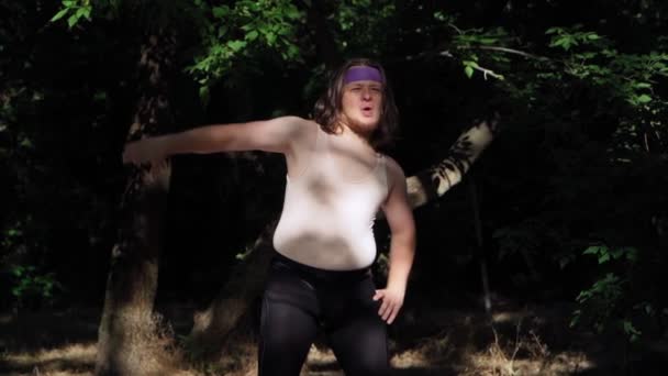 Playful Fat Man z długimi włosami w obcisłej koszuli i legginsy Czy fitness do odchudzania na zewnątrz. Zabawny facet z nadwagą Freak przedstawiający dziewczynę zaangażowaną w sport w lesie. Zwolniony ruch. - Materiał filmowy, wideo