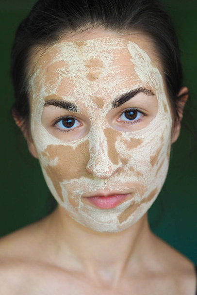 young brunette woman with a clay homemade mask on her face looks into the frame - Photo, Image