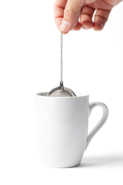 Metallische Teesieb Ei in einer menschlichen Hand Eintauchen und Einweichen in einer Tasse isoliert auf weiß - Foto, Bild