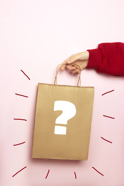 Жіноча рука в светрі тримає сумку для покупок на рожевому тлі. Екологічна торгова сумка з зображеним знаком питання в руках жінки. Творча ідея з графічним дизайном
 - Фото, зображення