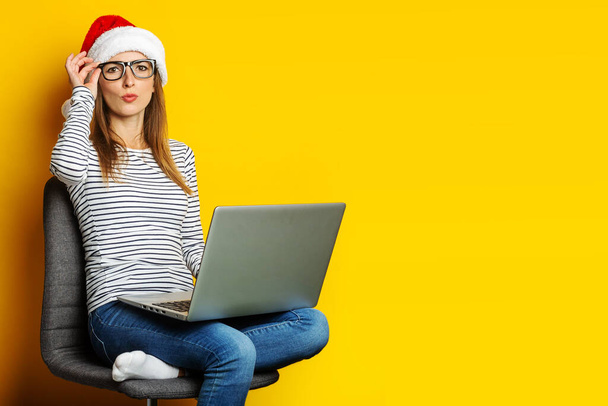 Młoda kobieta z laptopem, w kapeluszu Mikołaja i siedzi na krześle na żółtym tle. Sztandar. - Zdjęcie, obraz