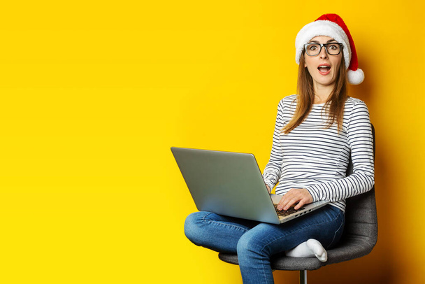 Jonge vrouw in een kerstmuts met een verrast gezicht zit op een stoel en houdt een laptop vast en is verbaasd over iets op een gele achtergrond. Banner. - Foto, afbeelding