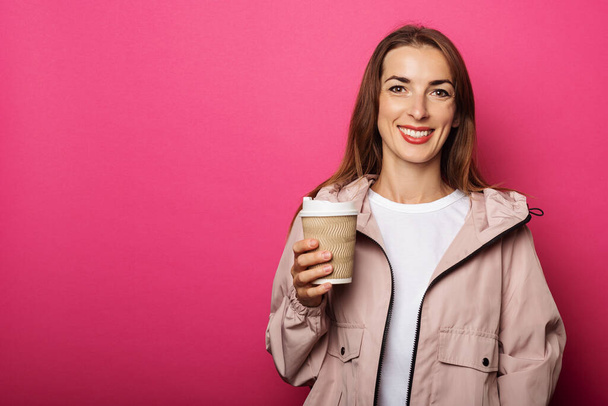 Mujer joven sonriendo y sosteniendo la taza de papel con café en la mano sobre fondo rosa. - Foto, imagen