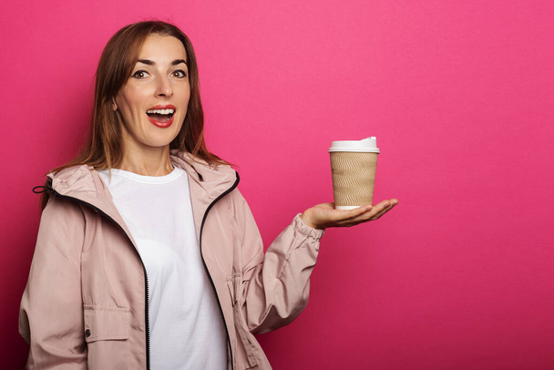 Young woman with surprised face holding paper cup with coffee in hand on pink background. - Photo, image