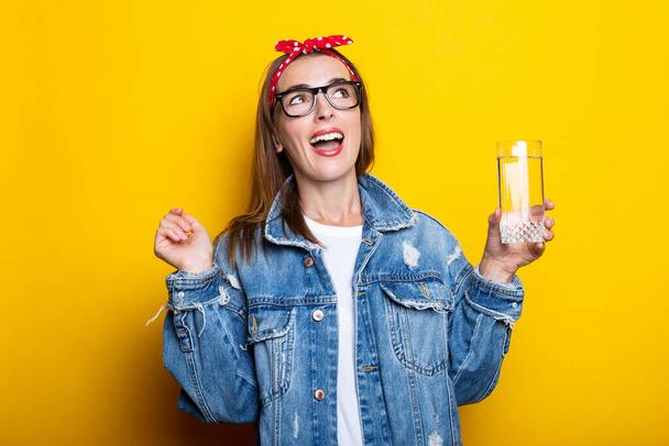 Jeune femme expressive en jeans, lunettes et bandeau tenant un verre d'eau propre sur fond jaune. - Photo, image