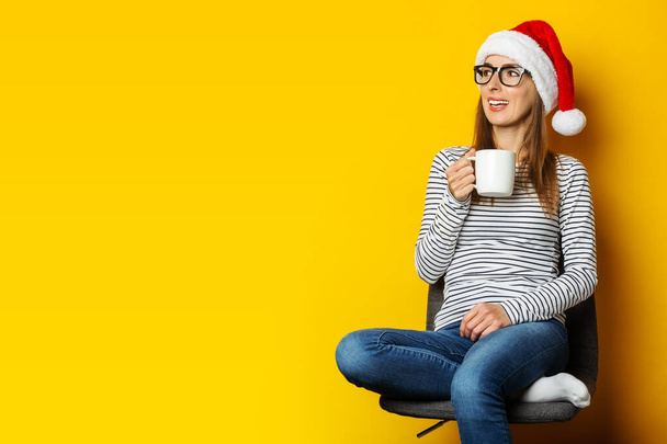 Young woman with surprised face in santa hat sits on a chair and holds a cup in her hands on a yellow background. - Foto, afbeelding
