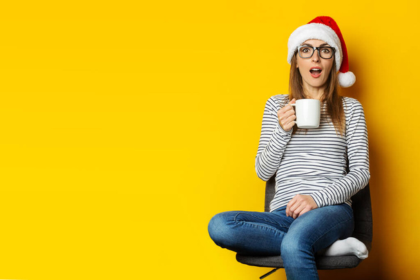 Junge Frau mit überraschtem Gesicht und Weihnachtsmannmütze sitzt auf einem Stuhl und hält auf gelbem Hintergrund eine Tasse in der Hand. - Foto, Bild