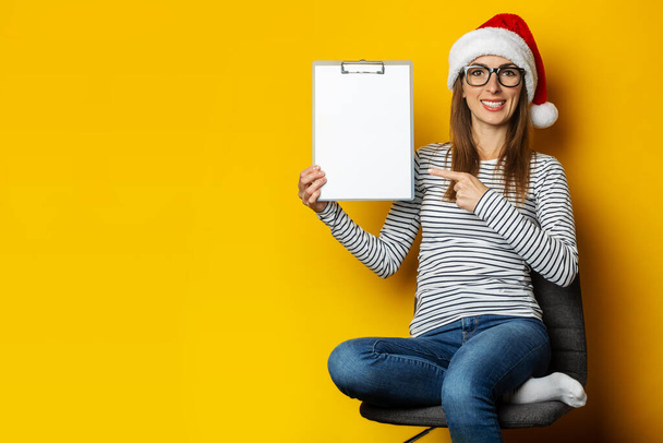 Giovane donna in un cappello di Babbo Natale si siede su una sedia e tiene una cartellina con un foglio di carta bianco su uno sfondo giallo. Bandiera. - Foto, immagini