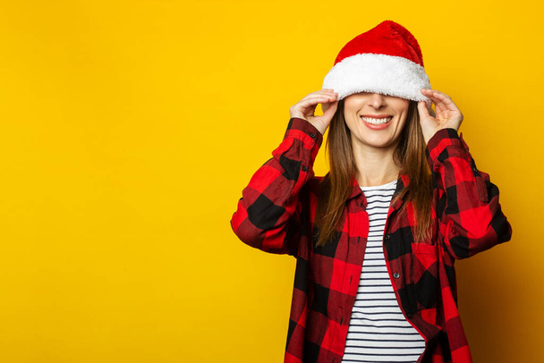 Young woman pulls santa hat and red plaid shirt over her eyes on yellow background. - Fotó, kép
