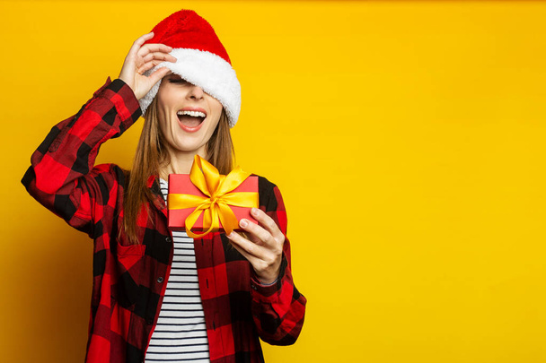 Junge Frau lugt unter Weihnachtsmannmütze und rot kariertem Hemd hervor und hält auf gelbem Hintergrund ein Geschenk in der Hand. - Foto, Bild