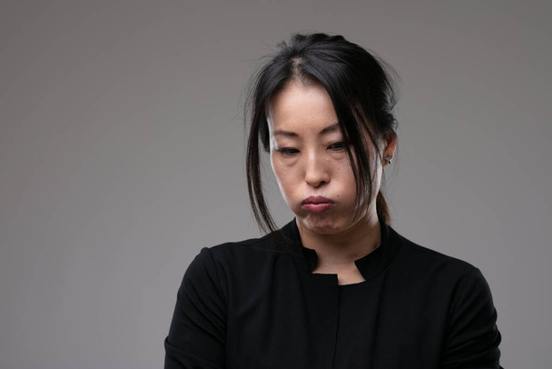 Kärttyisä murjottava aasialainen nainen murjottaa hänen huulensa ja katselee alas synkkä ilme yli harmaa studio tausta copyspace - Valokuva, kuva