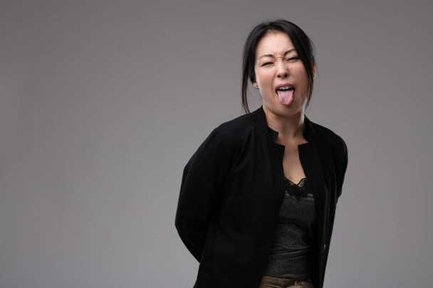 Freche, unhöfliche Asiatin streckt ihre Zunge heraus, während sie über einem grauen Studiohintergrund mit Kopierraum in die Kamera blickt - Foto, Bild