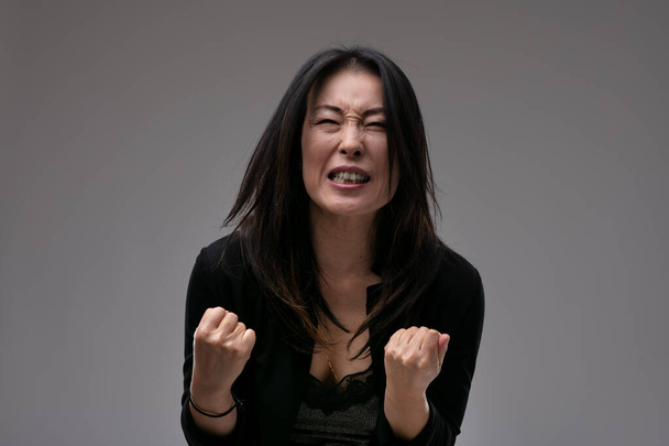 Mujer asiática emocionado molesto apretando los puños en la angustia como ella implora al espectador o es superado por la pérdida contra un fondo de estudio gris con copyspace - Foto, imagen
