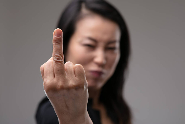 Rude aggressive Asian woman giving the middle finger in an offensive gesture with focus to her hand over grey - Photo, Image