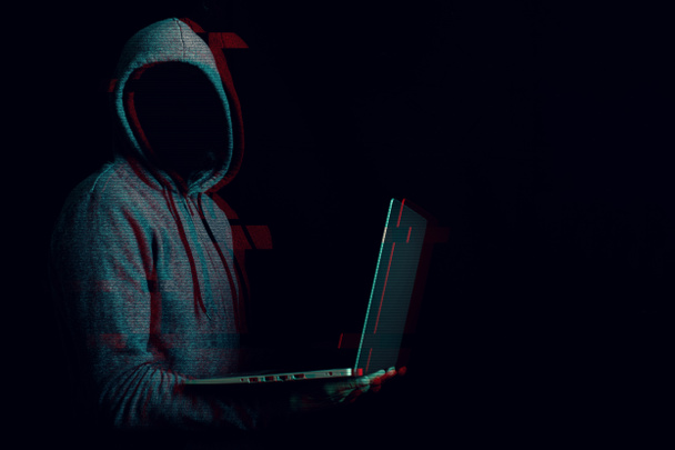 Man without a face in a hood holds a laptop in his hands on a dark background. Concept of cyber rogue, hacker. Banner. Added glitch effect. - Foto, afbeelding