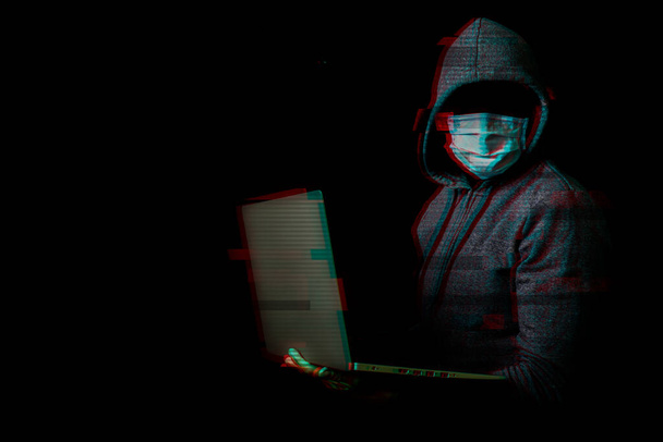 Mężczyzna w masce bez twarzy i kaptura trzyma laptopa w rękach na ciemnym tle. Koncepcja cyberłotra, hakerze. Sztandar. Dodano efekt usterki. - Zdjęcie, obraz
