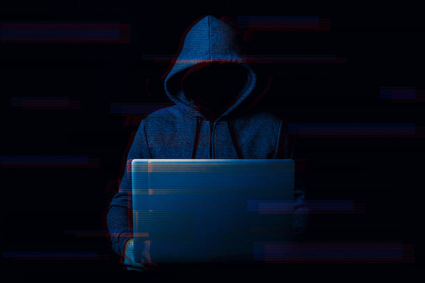 Człowiek bez twarzy w kapturze trzyma laptopa w rękach na ciemnym tle. Koncepcja cyberłotra, hakerze. Sztandar. Dodano efekt usterki. - Zdjęcie, obraz
