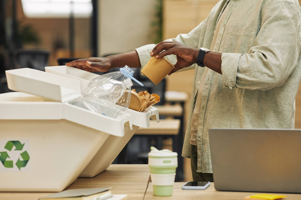 Portrait recadré d'un homme afro-américain méconnaissable mettant une tasse de papier dans une poubelle de tri des déchets dans un bureau, espace de copie - Photo, image