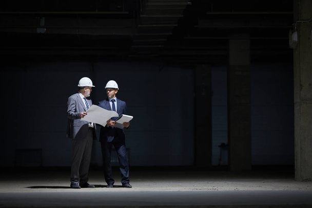 Dramaattinen täyspitkä muotokuva kaksi kypsää liikemiestä yllään kypärä ja tilalla suunnitelmia seistessään pimeässä työmaalla valaistu ankara valaistus, kopioi tilaa - Valokuva, kuva