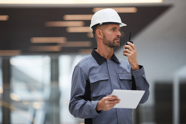 Taille omhoog portret van knappe volwassen werknemer spreken door walkie-talkie tijdens het toezicht op het werk op de bouwplaats of in industriële werkplaats, kopieerruimte - Foto, afbeelding
