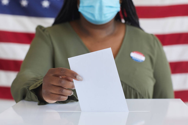 Primer plano de la mujer afroamericana usando máscara poniendo el boletín de votación en las urnas y mirando a la cámara mientras está de pie contra la bandera estadounidense el día de las elecciones, espacio para copiar - Foto, imagen