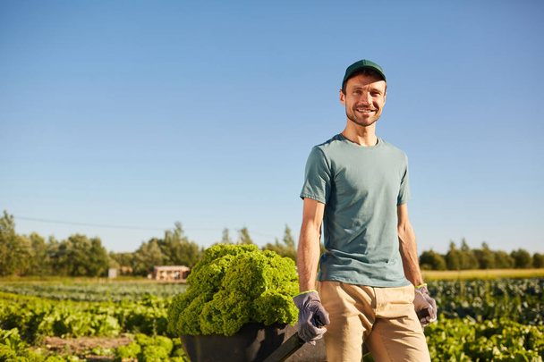 Портрет улыбающегося мужчины, который тянет тележку с богатым урожаем и смотрит в камеру, стоя на овощной плантации на открытом воздухе под солнечным светом, копирует пространство - Фото, изображение