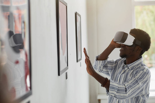 Ritratto laterale di un uomo afro-americano sorridente che indossa l'attrezzatura VR mentre si gode un'esperienza immersiva alla mostra di gallerie d'arte moderna, copia lo spazio - Foto, immagini