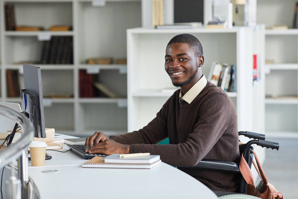 Portret niepełnosprawnego Afroamerykanina korzystającego z komputera i uśmiechającego się do kamery podczas studiów w bibliotece uczelni, przestrzeń kopiowania - Zdjęcie, obraz