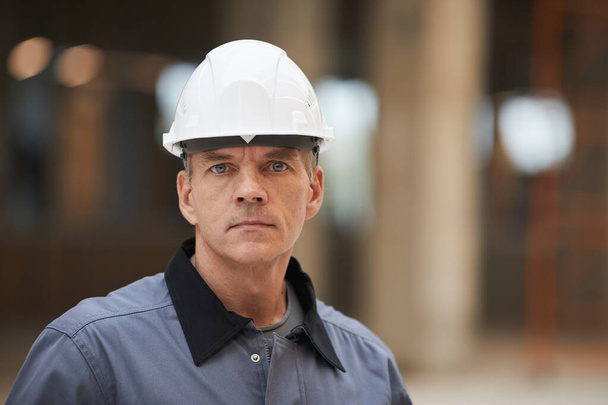 Close up portrait of mature worker wearing hardhat and looking at camera while standing at construction site or in industrial workshop, copy space - Photo, Image