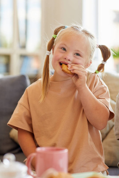 Vertikální portrét roztomilé dívky s Downovým syndromem jíst cookie při vychutnávání čaje s rodinou doma - Fotografie, Obrázek