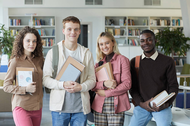 Többnemzetiségű diákcsoport portréja, akik kamerába néznek és mosolyognak, miközben a főiskolai könyvtárban állnak - Fotó, kép