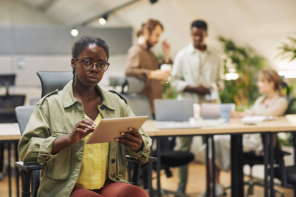 Portrait de jeune femme afro-américaine sérieuse regardant l'appareil photo tout en utilisant une tablette numérique assise dans un bureau moderne avec des personnes travaillant en arrière-plan, espace de copie - Photo, image