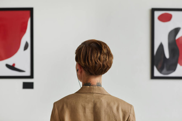 Graphic back view portrait of tattooed young woman looking at paintings and listening to audio guide at modern art gallery exhibition, copy space - Photo, Image