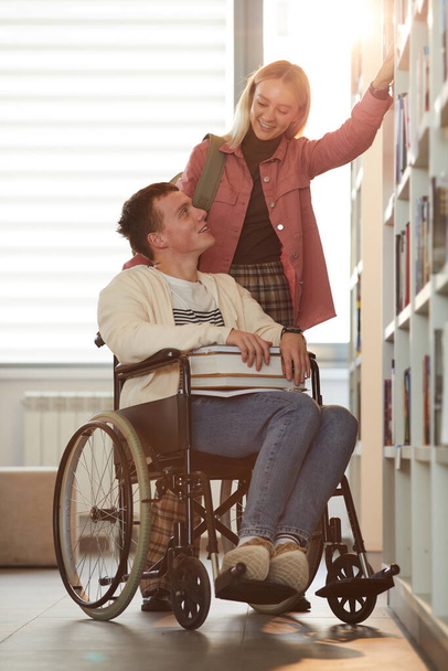 Κάθετη ολόσωμη προσωπογραφία του νεαρού άνδρα που χρησιμοποιεί αναπηρικό καροτσάκι στο σχολείο με γυναίκα φίλη που τον βοηθάει στη βιβλιοθήκη φωτισμένη από το φως του ήλιου - Φωτογραφία, εικόνα