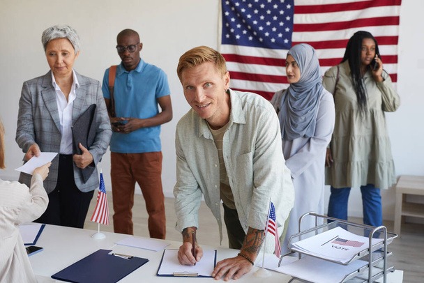 Многонациональная группа людей, регистрирующаяся на избирательном участке, украшенном американскими флагами в день выборов, сосредоточена на улыбающемся человеке, подписывающем избирательные бюллетени и смотрящем в камеру, копирующем пространство - Фото, изображение
