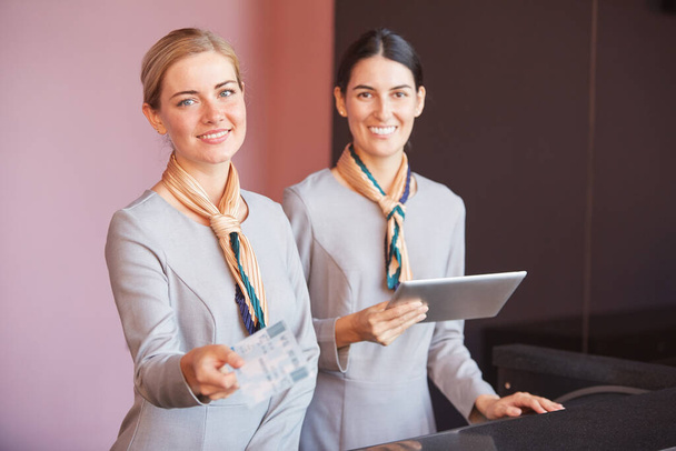 Portrait of two smiling flight attendants handing tickets to passenger while standing at check in desk in airport, copy space - Photo, Image